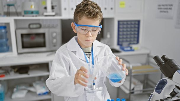 Schattige blonde kind wetenschapper serieus bezig met een chemie-experiment, gieten vloeistof in reageerbuis tijdens het werken in het lab - Foto, afbeelding
