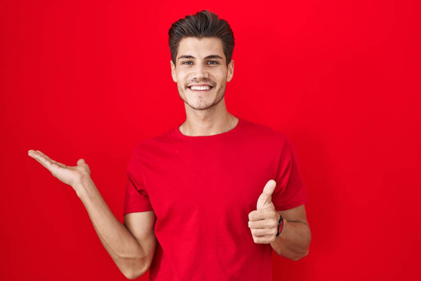 Mladý hispánec, který stojí nad červeným pozadím a ukazuje dlaň a dělá dobře gesto s palci nahoru, usmívá se šťastný a veselý  - Fotografie, Obrázek