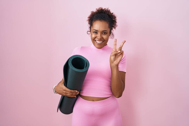 Jeune femme hispanique aux cheveux bouclés tenant tapis de yoga sur fond rose souriant avec un sourire heureux clin d'oeil à la caméra faisant signe de victoire avec les doigts. numéro deux.  - Photo, image