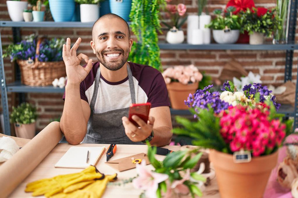 Іспаномовний чоловік з бородою, що працює у квітковому магазині зі смартфоном, робить знак ок з пальцями, усміхнений дружній жест відмінний символ  - Фото, зображення
