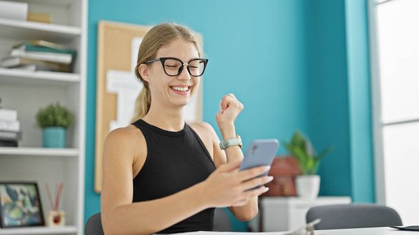 Jonge blonde vrouw zakenman met behulp van smartphone met winnaar gebaar op kantoor - Foto, afbeelding