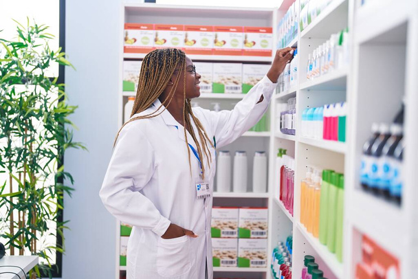 Africano americano mulher farmacêutico sorrindo confiante segurando desodorante de estantes na farmácia - Foto, Imagem