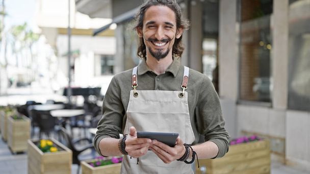 Joven camarero hispano sonriendo confiado usando touchpad en la terraza de la cafetería - Foto, Imagen