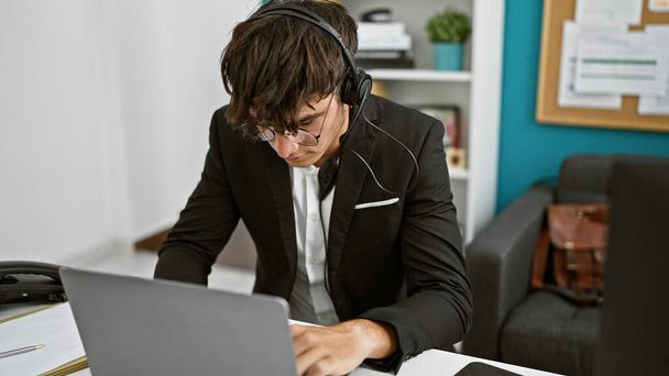 Νέος, όμορφος Ισπανόφωνος έφηβος που τρέχει στο γραφείο, φοράει ακουστικά, ασχολείται με επαγγελματική δουλειά, με λέιζερ επικεντρωμένο στην επιτυχία του laptop. - Φωτογραφία, εικόνα