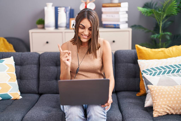 Young hispanic woman working from home wearing operator headset smiling happy pointing with hand and finger to the side  - Photo, Image