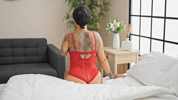 Mulher hispânica com braço amputado vestindo lingerie sentada na cama para trás no quarto - Foto, Imagem