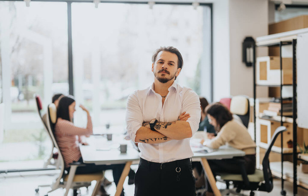 Татуированный мужчина-предприниматель излучает уверенность, стоя со скрещенными руками в хорошо освещенной обстановке офиса, в то время как коллеги занимаются работой позади него.. - Фото, изображение