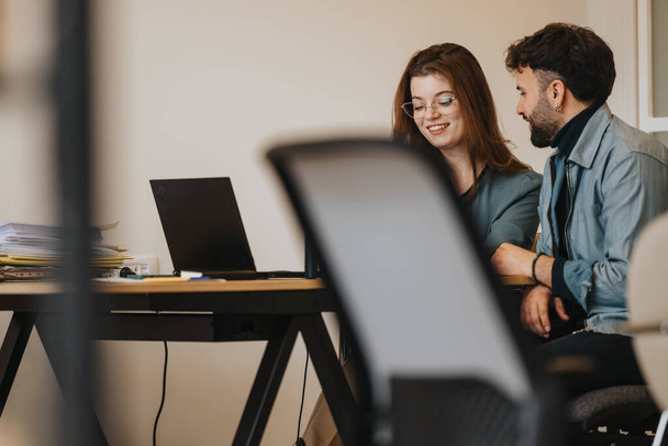 Twee zakelijke professionals die in gesprek gaan aan een bureau met een laptop, in een moderne kantooromgeving. - Foto, afbeelding