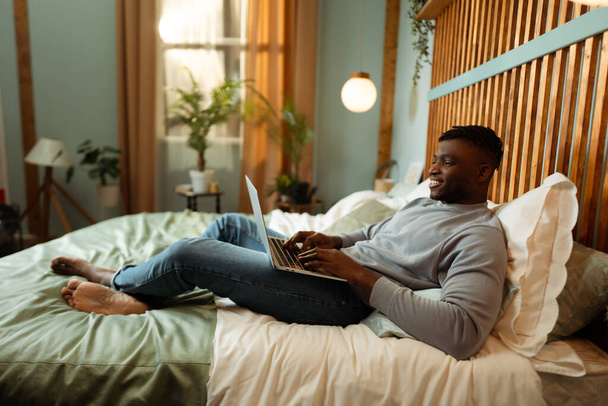 Mutlu Afro-Amerikan bir adamın portresi, dizüstü bilgisayar kullanan, evde yatakta uzanan, rahatlatıcı, metin yazarı. Uzak iş kavramı, online teknoloji - Fotoğraf, Görsel