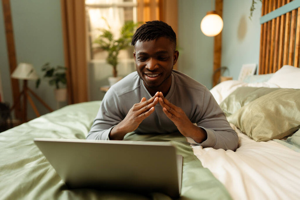 Retrato de homem afro-americano sorridente, hipster tendo videochamada, gestos, mostrando gestos de linguagem gestual, explicando algo enquanto estava deitado na cama em casa. Conceito de comunicação - Foto, Imagem