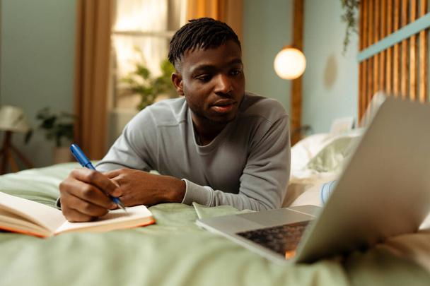 Retrato de um jovem afro-americano pensativo, estudante usando laptop, tomando notas, preparando-se para o exame enquanto estava deitado na cama em casa. Conceito de educação online, tecnologia - Foto, Imagem