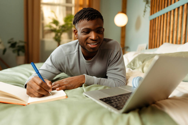 Retrato de homem afro-americano atraente sorridente, estudante usando laptop, tomando notas, se preparando para o exame enquanto deitado na cama em casa. Conceito de educação online, tecnologia - Foto, Imagem