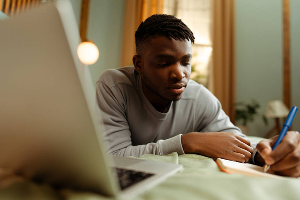 Retrato de homem afro-americano bonito e pensativo deitado na cama em casa, estudando online, usando laptop. Escritor jovem segurando caneta e tomando notas. Conceito de educação online, aprendizagem de línguas - Foto, Imagem