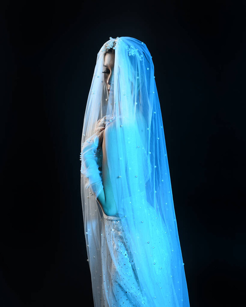 Close up portrait of beautiful model wearing white wedding gown and ghostly flowing veil like a shroud. Moody cinematic lighting, isolated on a dark studio background. - Photo, Image