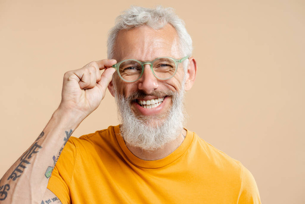 Lächelnder erfolgreicher älterer Mann, mit stylischer Brille und lässig gelbem T-Shirt, in die Kamera blickend, isoliert auf beigem Hintergrund stehend. Vision-Konzept - Foto, Bild