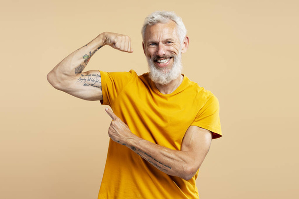 Porträt eines grauhaarigen Mannes mittleren Alters, der lässig gelbes T-Shirt trägt und Bizeps zeigt, mit dem Finger auf tätowierte Hand zeigt, in die Kamera blickt und isoliert auf beigem Hintergrund steht. Sportkonzept - Foto, Bild
