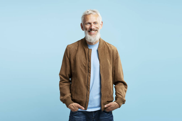 Портрет усміхненого бородатого сірого волосистого чоловіка в стильній повсякденній коричневій куртці, який дивиться на камеру, стоїть ізольовано на бежевому фоні. Концепція шопінгу, реклама - Фото, зображення