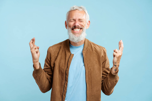 Gülümseyen gri sakallı olgun bir adamın portresi şık bir ceket giyen, parmakları çapraz, gözleri kapalı mavi arka planda duran. Umut kavramı - Fotoğraf, Görsel