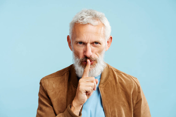 Portrét atraktivního šedovlasého muže v nedbale hnědé bundě s nápisem mlčení, který se dívá na kameru stojící izolovaně na modrém pozadí. Tajný koncept - Fotografie, Obrázek