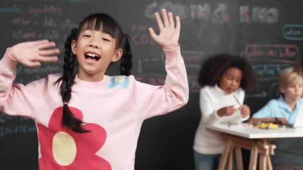 アジアの少女は笑顔とカメラに波,友人が働いているか,エンジニアリングコードを学んでいる間,またはSTEMの技術教室でプロンプト. カメラを見て,子供がノートパソコンを使って挨拶をする. エラー. - 映像、動画