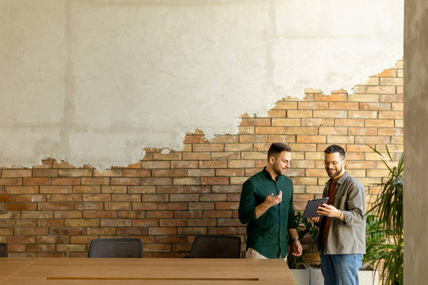 Dois profissionais estão envolvidos em uma conversa focada enquanto mantêm o tablet digital em um espaço de escritório contemporâneo, apresentando tijolos expostos rústicos e luz natural quente. - Foto, Imagem