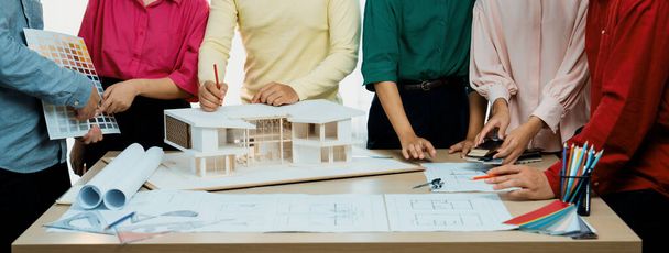Przycięty obraz profesjonalnego zespołu architektów burza mózgów o wyborze koloru domu, podczas gdy architekt pisze na dokumencie projektu przy stole umieszczone z planem i modelu domu. Różnorodność. - Zdjęcie, obraz