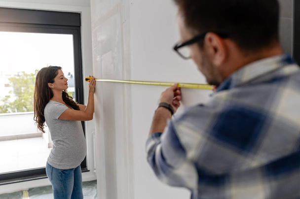 Una pareja joven, hermosa y alegre, compuesta por un hombre y su esposa embarazada, están midiendo las paredes, como parte de su nuevo proyecto de renovación del hogar - Foto, Imagen