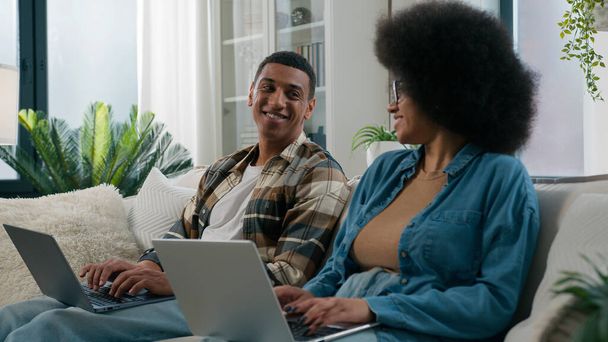 Africká americká žena pracovat s notebookem muž počítač pracovní pár sedět v řadě dva nezávislí pracovníci s počítači, kteří spolupracují doma závislý rodina při pohledu na sebe smějící se hraní hry on-line na pohovce - Fotografie, Obrázek