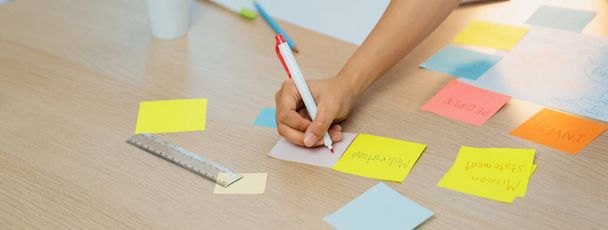 Close-up beeld van jonge creatieve zakenvrouw schrijft op kleurrijke plakkerige notitie en plaatst het op brainstorming mindmap in de vergaderzaal. Creatief en teamwork concept. Focus op de hand. Afgerond. - Foto, afbeelding
