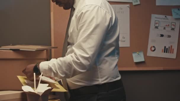 Medium shot van Black mannelijke executive stand-by kurkboard met business charts en diagrammen op, het analyseren van de voortgang van het bedrijf en het eten van afhaalmaaltijden met eetstokjes tijdens het werken 's nachts - Video