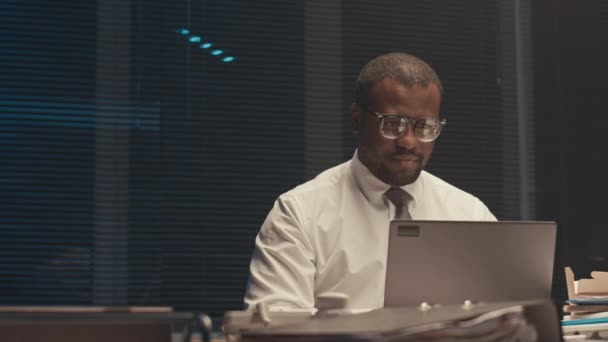 Περίμενε έναν πολυάσχολο Αφροαμερικανό επιχειρηματία με γυαλιά που κοιτάζει το λάπτοπ και κρατάει σημειώσεις για τα επαγγελματικά έγγραφα ενώ δουλεύει υπερωρίες στο γραφείο αργά το βράδυ. - Πλάνα, βίντεο
