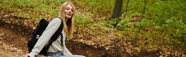 Улыбающаяся расслабленная блондинка-туристка в свитере и рюкзаке сидит на скале в лесу, баннер - Фото, изображение