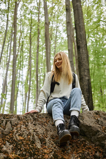 Smiling hiking blonde woman wearing sweater and backpack sitting on a rock in woods - Fotó, kép