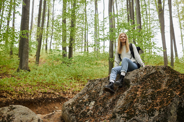 Active young blonde woman hiker sitting on a rock on a backpacking trip in peaceful forest - 写真・画像