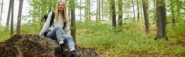 Smiling pretty blonde woman hiker sitting on a rock in forest and looking away, banner - Fotó, kép