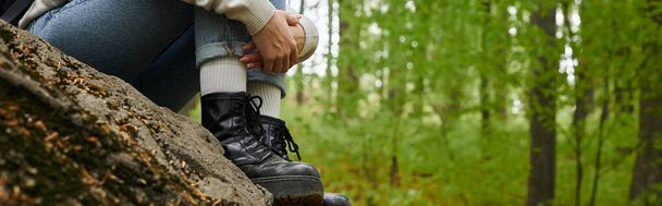Cropped photo of female hiker hugging her legs wearing jeans and hiking boots in forest, banner - Фото, зображення