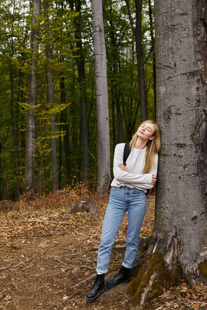 Full height portret van blonde vrouw in wandelen outfit in het bos leunend op boom met ogen dicht - Foto, afbeelding