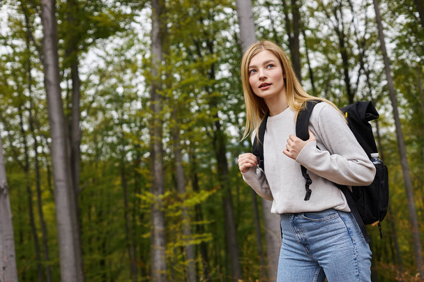 Curiosa donna bionda che tiene lo zaino, indossa maglione e jeans, guarda attraverso i boschi - Foto, immagini