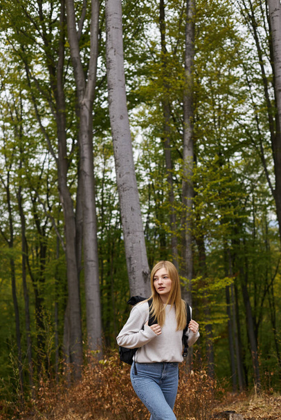 Enthusiastic and blonde girl in green forest wearing sweater and jeans, exploring scenic view - Zdjęcie, obraz