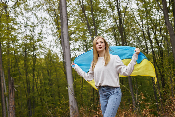 Wspierająca niezależna blondynka stojąca w lesie z ukraińską flagą machającą nią - Zdjęcie, obraz