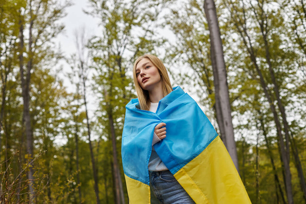 Mulher patriótica de pé na floresta cobrindo corpo com bandeira ucraniana e olhando na frente dela - Foto, Imagem
