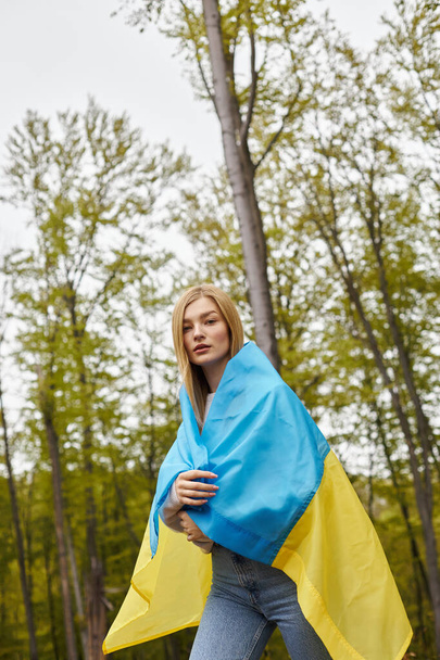Patriotyczna blondynka dumnie trzyma narodową ukraińską flagę w lesie i patrzy w kamerę - Zdjęcie, obraz