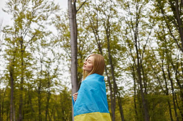 Inspired blonde girl holding the Ukrainian flag over her body in forest with closed eyes - Photo, Image