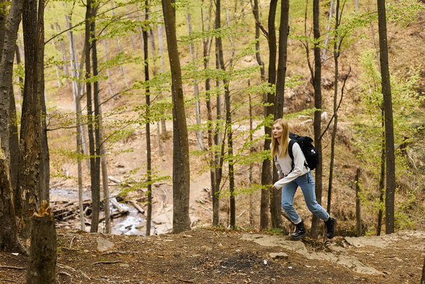 Enthusiastic blond girl in forest wearing sweater and jeans, running and exploring scenic view - Fotoğraf, Görsel
