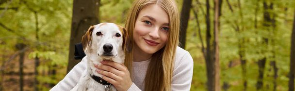 Smiling blonde woman hugging her dog, while having a halt on forest trip, looking at camera, banner - Фото, изображение