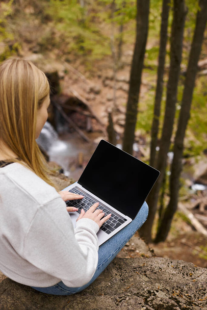 mujer rubia teniendo viaje de mochilero y descansando en la roca en el bosque, trabajando en el ordenador portátil - Foto, Imagen