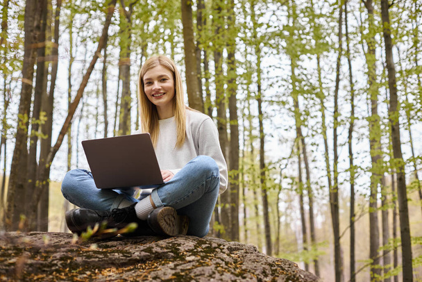 Smiling bright woman on halt in woods, sitting on boulder with laptop in lotus position - Zdjęcie, obraz