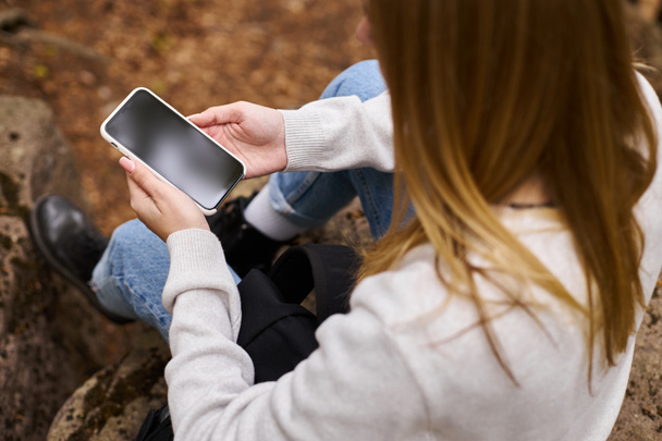 Zugeschnittenes Bild einer Frau, die ihr Handy in der Hand hält, in einer Waldlandschaft sitzt und SMS schreibt - Foto, Bild