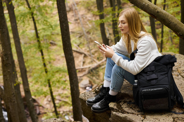Vue latérale de la femme tenant le téléphone tout en se reposant dans la forêt. Concept de randonnée et de tourisme - Photo, image