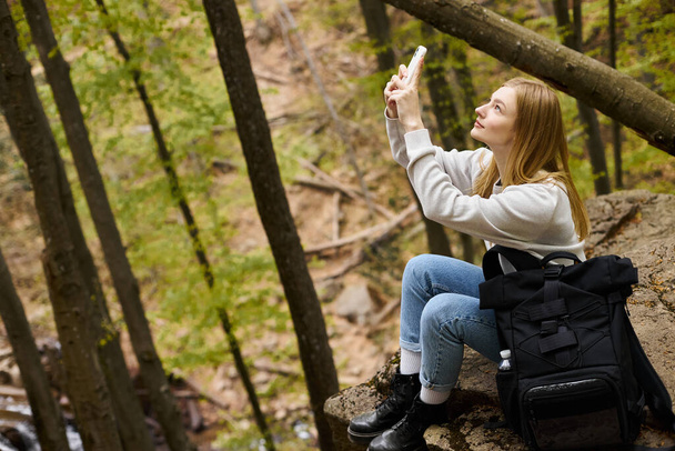 Вид сбоку на блондинку, фотографирующую пейзаж в лесу во время похода - Фото, изображение